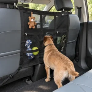 A dog peeking through the circular opening of a black mesh car barrier, with a hanging plush toy and pet travel supplies in the storage pockets.